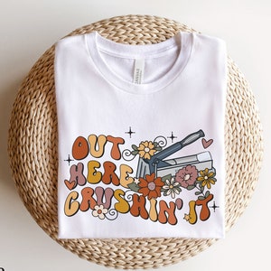 May include: White t-shirt with the text "Out here crushin' it" in colorful, retro-style lettering. The text is surrounded by flowers and a stapler.