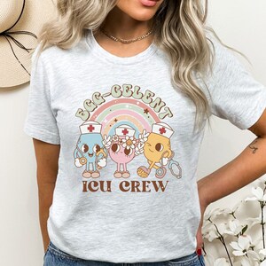 May include: A gray t-shirt with a rainbow and three cartoon characters wearing nurse hats. The text "Egg-Celent ICU Crew" is printed on the shirt.