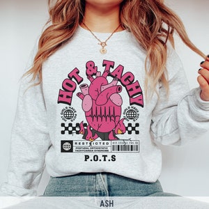 May include: Grey sweatshirt with a pink heart graphic and the text "HOT & TACHY" in black. The heart has a black EKG line and flames. The text "GIRLS HAVE POTS" is in a circle around the heart. Below the heart is a black and white barcode with the text "RESTRICTED POSTURAL ORTHOSTATIC TACHYCARDIA SYNDROME P.O.T.S."