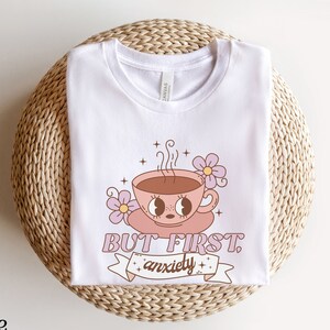 May include: White t-shirt with a graphic of a pink coffee cup with eyes and the text "But First, Anxiety".