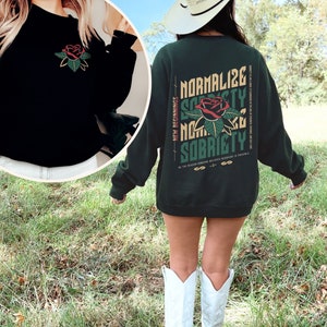 May include: A dark green sweatshirt with a rose graphic and the text "Normalize Sobriety" on the back. The text is in a white, black, and yellow font. The rose is red with green leaves. The sweatshirt is being worn by a person with a white cowboy hat.