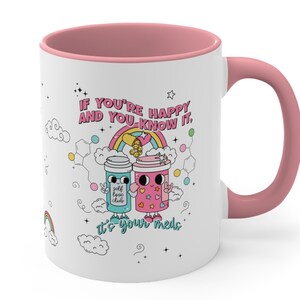 May include: White ceramic mug with a pink rim and handle. The mug features a colourful illustration of two cartoon coffee cups with the text "IF YOU'RE HAPPY AND YOU KNOW IT. It's your meds."