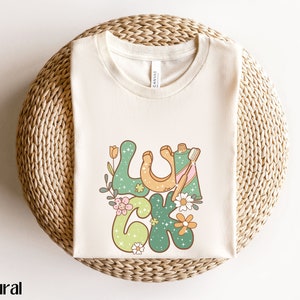 Peut inclure: T-shirt crème avec le mot "LUCK" dans une police rétro, orné de fleurs et d'un fer à cheval. Le t-shirt a un col rond et est fait d'une matière douce. Le mot "natural" est imprimé en bas à gauche.