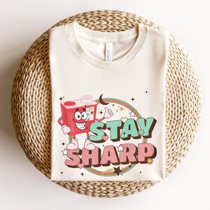 May include: A white t-shirt with a cartoon illustration of a red box with a face and arms. The box is holding a thumbs up and the text "STAY SHARP" is written in green and pink letters.