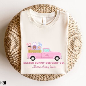 May include: Cream-colored t-shirt featuring a pink truck with the text "Easter Bunny Delivery Co." and "Mother Baby Unit." The truck bed holds three cartoon bunnies. The word "natural" is printed at the bottom left.