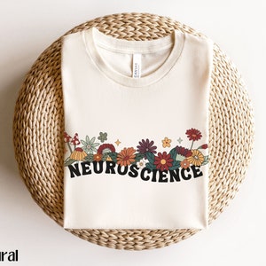 May include: A natural colored t-shirt with a floral design and the word "NEUROSCIENCE" in black lettering.