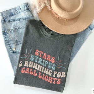 May include: A dark gray t-shirt with a retro-style graphic that says "Stars Stripes & Running for Call Lights" in red, blue, and white. The graphic includes fireworks.