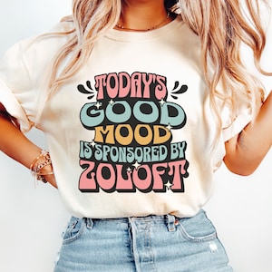May include: A white t-shirt with the text "Today's Good Mood Is Sponsored By Zoloft" in colorful retro-style lettering.