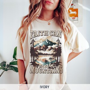 May include: A cream coloured t-shirt with a graphic of a mountain range and a lake. The text "Faith Can Move Mountains" is printed on the shirt.