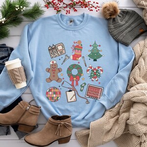 May include: Light blue sweatshirt with a Christmas-themed design featuring a gingerbread person, a stocking, a Christmas tree, a wreath, candy canes, a stethoscope, a bandage, a disco ball, a medical chart, and an EKG monitor.