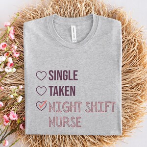 May include: Gray t-shirt with a pink heart graphic and the text "Single, Taken, Night Shift Nurse" in purple.