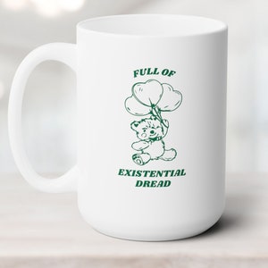 May include: White ceramic mug with a green outline of a teddy bear holding balloons that say "Full of Existential Dread".