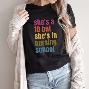 May include: Black t-shirt with the text "she's a 10 but she's in nursing school" in colorful letters. The shirt is worn with a cream-colored cardigan and blue jeans. The word "BLACK" is printed in the upper right corner.