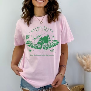 May include: Pink t-shirt with a green graphic of the Earth and the text "Being Alive is the Special Occasion" with butterflies and stars around it. The text below the graphic says "Find the beauty. Enjoy the journey."