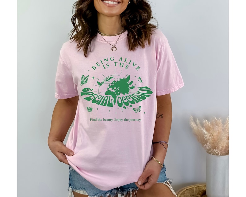May include: Pink t-shirt with a green graphic of the Earth and the text "Being Alive is the Special Occasion" with butterflies and stars around it. The text below the graphic says "Find the beauty. Enjoy the journey."