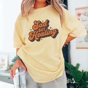Puede incluir: Una camiseta amarilla clara con el texto "Sad but Healing" en letras naranjas y negras. La camiseta tiene un corte holgado y parece estar hecha de un material suave. El dise&ntilde;o tiene una est&eacute;tica retro.