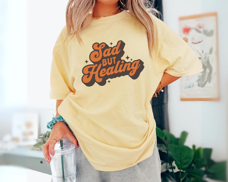 Puede incluir: Una camiseta amarilla clara con el texto "Sad but Healing" en letras naranjas y negras. La camiseta tiene un corte holgado y parece estar hecha de un material suave. El dise&ntilde;o tiene una est&eacute;tica retro.
