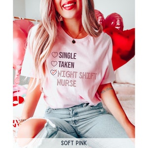 May include: A pink t-shirt with the text "Single, Taken, Night Shift Nurse" with a checkmark next to the last option. The shirt is worn by a person with blonde hair and blue jeans.
