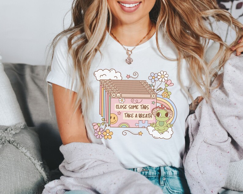 May include: White t-shirt with a pink and green graphic of a frog sitting on a cloud with the text "Close some tabs Take a breath".