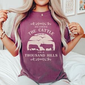 May include: A purple t-shirt with a graphic design that says "He owns the cattle on a thousand hills, Psalm 50". The graphic also includes a silhouette of two cows.
