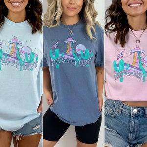 May include: Three t-shirts in light blue, blue-grey, and pink. Each shirt features a colourful graphic with a UFO, rocket ship, cactus, and the text "LET'S GET LOST". The design has a retro, psychedelic vibe.