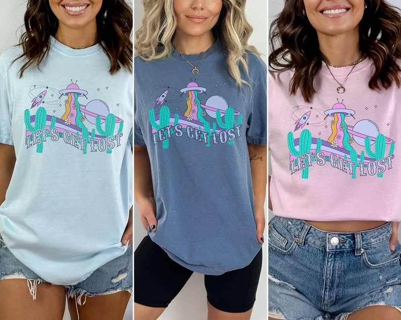 May include: Three t-shirts in light blue, blue-grey, and pink. Each shirt features a colourful graphic with a UFO, rocket ship, cactus, and the text "LET'S GET LOST". The design has a retro, psychedelic vibe.