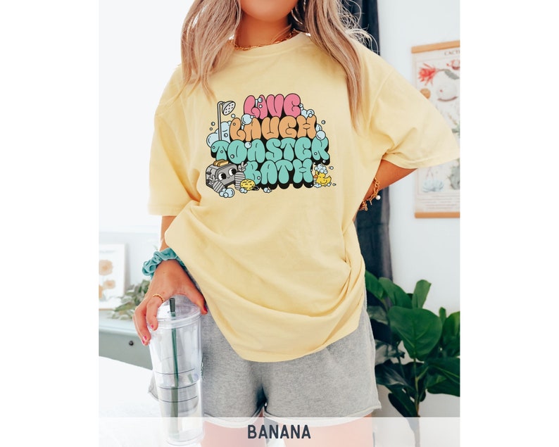 Pu&ograve; includere: Una maglietta gialla con una stampa grafica che dice "Live Laugh Take a Toaster Bath". La grafica &egrave; in stile retr&ograve; con lettere a bolle colorate.