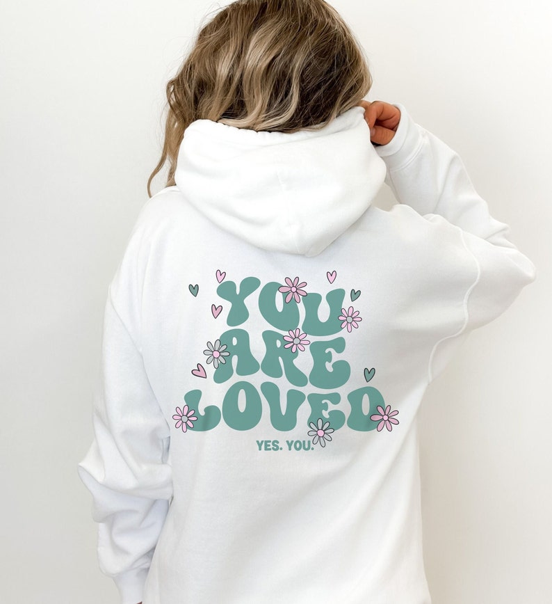 以下が含まれることがあります： 白のパーカー。 ターコイズ色の「YOU ARE LOVED」の文字とピンクの花とハートが描かれています。「YES. YOU.」の文字が下にあります。 パーカーには、ドローストリングフードと長袖が付いています。