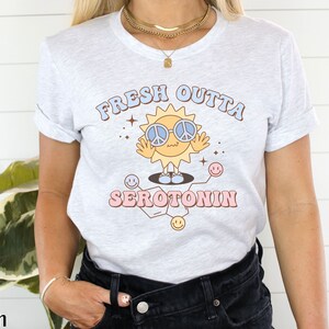 Op de afbeelding: Lichtgrijs t-shirt met de tekst "FRESH OUTTA SEROTONIN" en een cartoon zon met een peace-teken zonnebril. De zon staat op een moleculaire structuur met smiley gezichten. Het shirt heeft opgerolde mouwen.