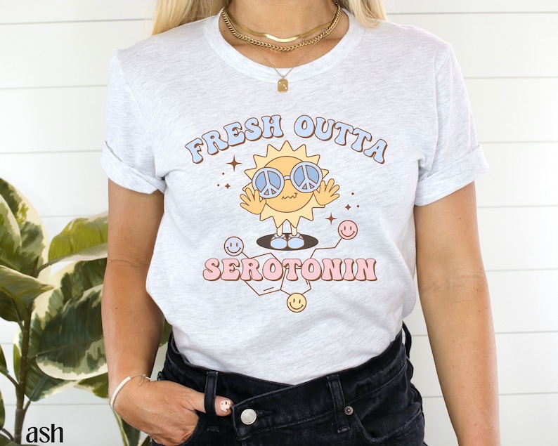 Op de afbeelding: Lichtgrijs t-shirt met de tekst "FRESH OUTTA SEROTONIN" en een cartoon zon met een peace-teken zonnebril. De zon staat op een moleculaire structuur met smiley gezichten. Het shirt heeft opgerolde mouwen.