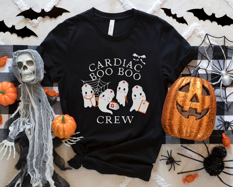 May include: Black t-shirt with a white graphic of three ghosts wearing nurse hats and holding medical equipment. The text "Cardiac Boo Boo Crew" is printed on the shirt.