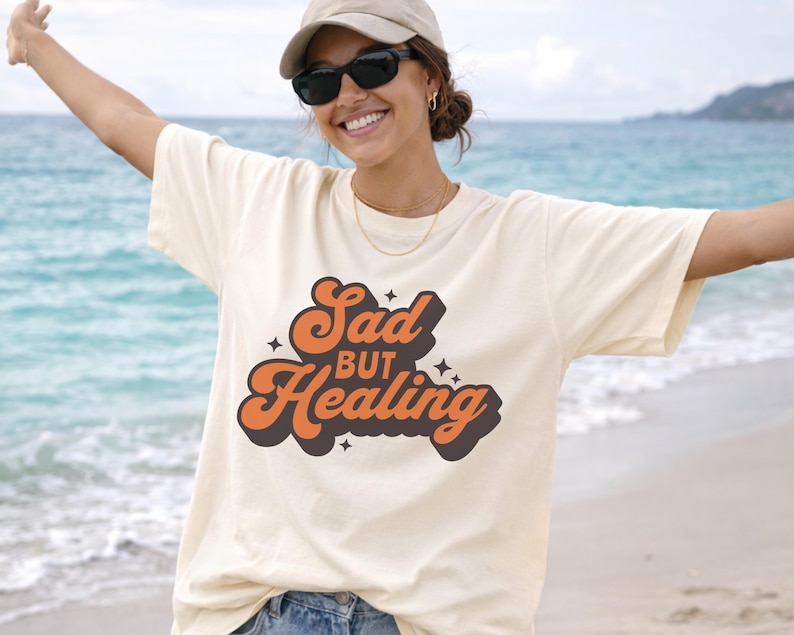 Puede incluir: Camiseta color crema con el texto "Sad but Healing" en fuente retro naranja y marr&oacute;n. El dise&ntilde;o incluye estrellas. Una prenda casual para uso diario.