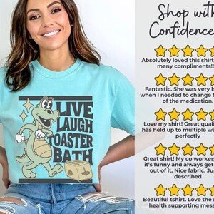 May include: A light blue t-shirt with a cartoon dinosaur graphic and the words "LIVE LAUGH TOASTER BATH". The dinosaur is green and beige, and the toaster is beige. The shirt also has customer reviews.