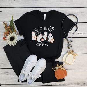 May include: Black t-shirt with a white graphic of ghosts dressed as nurses. The text "BOO BOO CREW" is printed on the shirt.