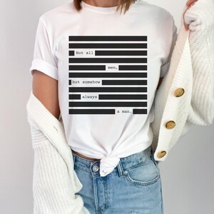May include: White t-shirt with a black and white graphic design. The design features the text "Not all men, but somehow always a man." The shirt is a classic crew neck style.