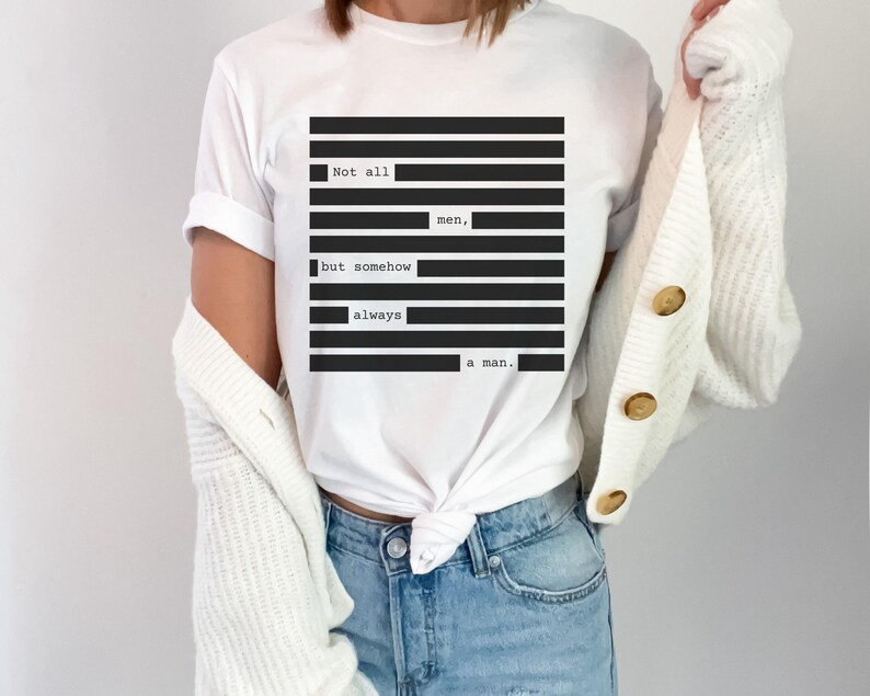 May include: White t-shirt with a black and white graphic design. The design features the text "Not all men, but somehow always a man." The shirt is a classic crew neck style.