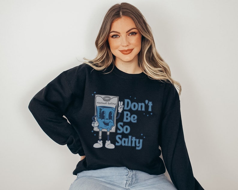 Op de afbeelding: Zwart sweatshirt met een blauwe en witte cartoonillustratie van een fysiologisch zoutoplossingfles met de tekst "Don't Be So Salty".