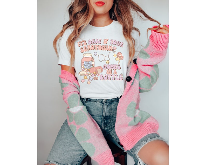 May include: White t-shirt with a pink and white graphic that says "It's okay if your serotonin comes in a bottle". The graphic includes a bottle labeled "Self Love Club" with a pink and white chemical structure and other pink and white illustrations.