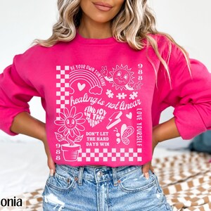 May include: Pink sweatshirt with a white graphic print featuring a sunflower, a rainbow, a sun, a butterfly, and the text "BE YOUR OWN SUNSHINE", "healing is not linear", "FIND JOY IN THE JOURNEY", "DON'T LET THE HARD DAYS WIN", "LOVE YOURSELF", and "988".