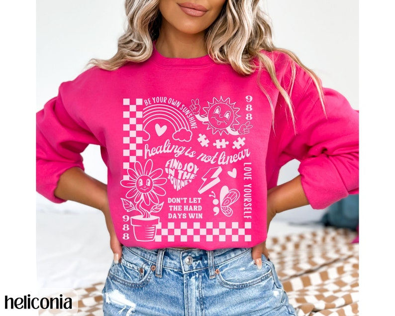 May include: Pink sweatshirt with a white graphic print featuring a sunflower, a rainbow, a sun, a butterfly, and the text "BE YOUR OWN SUNSHINE", "healing is not linear", "FIND JOY IN THE JOURNEY", "DON'T LET THE HARD DAYS WIN", "LOVE YOURSELF", and "988".