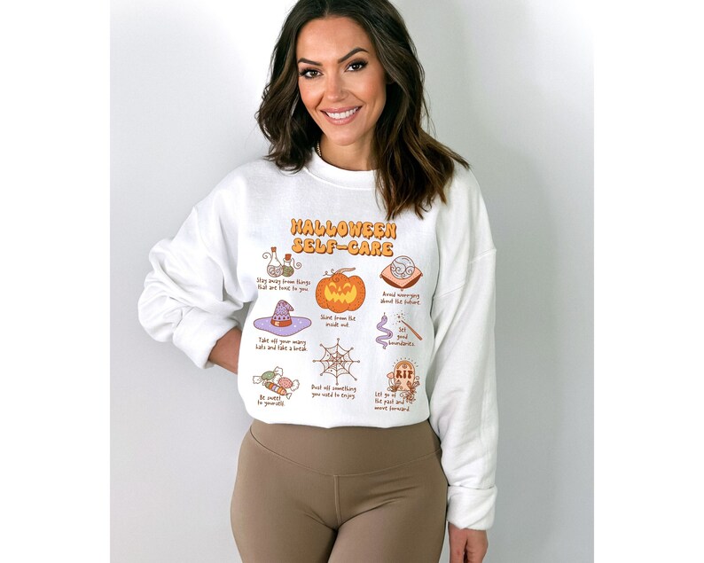 May include: White sweatshirt with a graphic print of a pumpkin, witch hat, spider web, and other Halloween-themed illustrations. The text "Halloween Self-Care" is printed in orange and yellow letters.