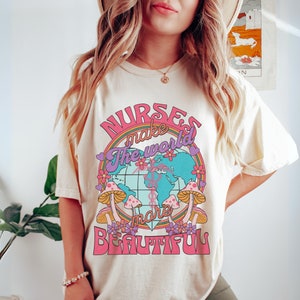 Puede incluir: Una camiseta blanca con un dise&ntilde;o gr&aacute;fico rosa, morado y azul que dice "Las enfermeras hacen que el mundo sea m&aacute;s hermoso". El gr&aacute;fico presenta un globo terr&aacute;queo con flores y hongos.