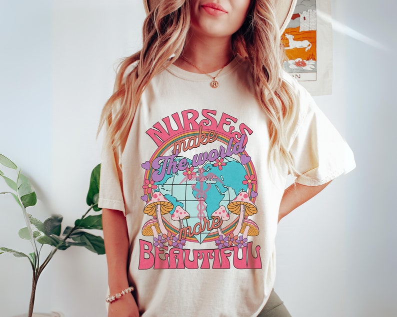 Puede incluir: Una camiseta blanca con un dise&ntilde;o gr&aacute;fico rosa, morado y azul que dice "Las enfermeras hacen que el mundo sea m&aacute;s hermoso". El gr&aacute;fico presenta un globo terr&aacute;queo con flores y hongos.