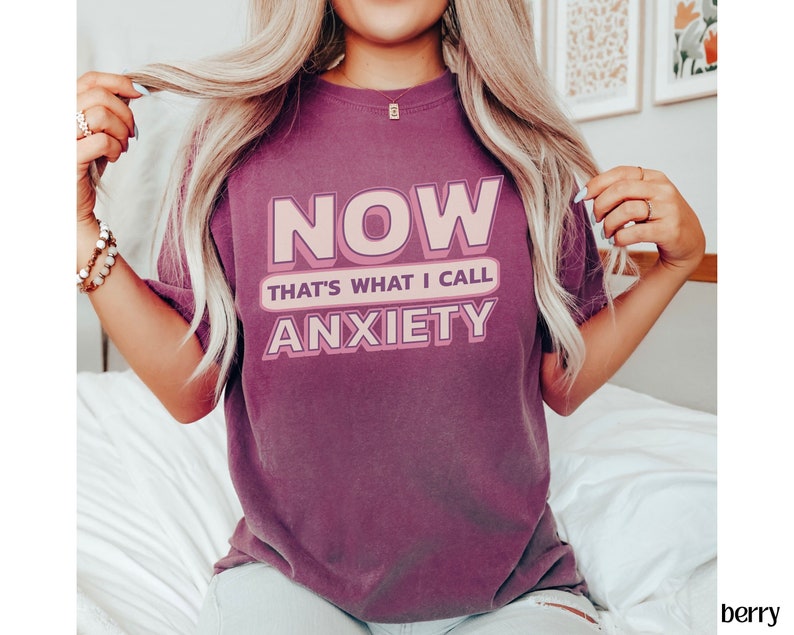 May include: Berry-colored t-shirt with the text "NOW THAT'S WHAT I CALL ANXIETY" in a pink and white design. The shirt is a crew neck style and appears to be made of a soft, comfortable material.