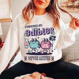 May include: White sweatshirt with a graphic of two cartoon gummies with the text "Powered by: Edibles" and "Garden Gummies to the Rescue".