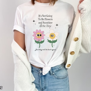 May include: White t-shirt with a graphic of two cartoon flowers with faces. The text on the shirt reads "It's Not Going To Be Flowers and Sunshine All the Time. But they will be back again."