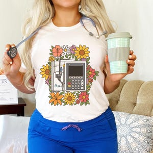 May include: White t-shirt featuring a medical device graphic surrounded by colorful flowers. The shirt has a crew neck and is paired with blue scrubs. A stethoscope and a reusable coffee cup are also visible.