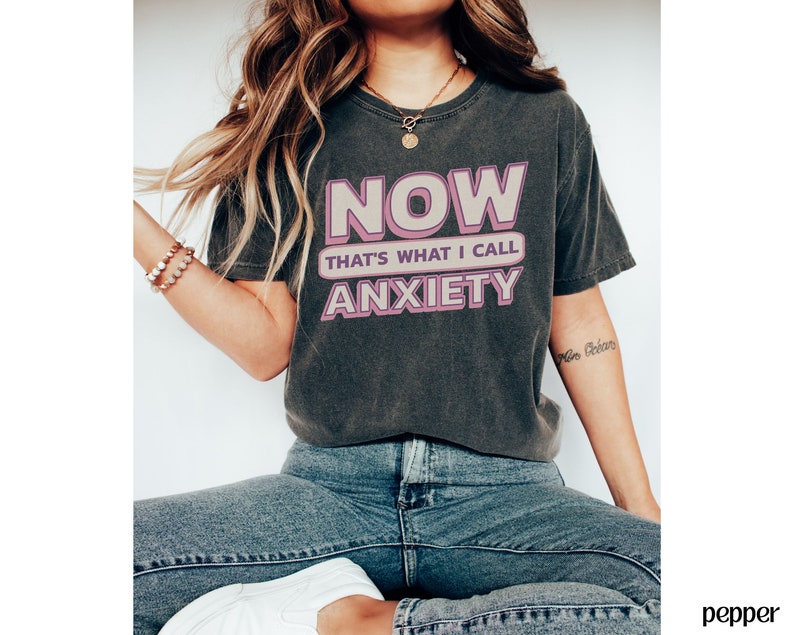 May include: Dark gray t-shirt with the text "NOW THAT'S WHAT I CALL ANXIETY" in pink and white. The shirt has a vintage, worn look. The model is wearing blue jeans.
