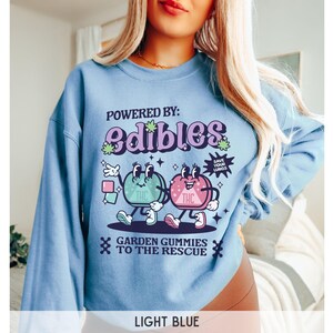 May include: Light blue sweatshirt with a graphic of two cartoon gummies with the text "POWERED BY: edibles" and "GARDEN GUMMIES TO THE RESCUE".