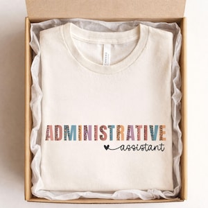 May include: A cream-coloured t-shirt featuring the text "ADMINISTRATIVE assistant" in colourful block letters and a small heart. The shirt is presented folded inside a cardboard gift box.